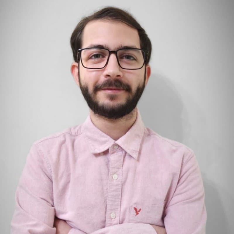 Smiling man with a beard and glasses wearing a pink shirt against a neutral background.